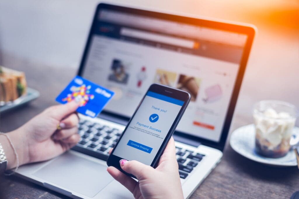 Person holding a credit card and smartphone with a "Payment Success" message displayed, in front of a laptop with an online shopping website open.